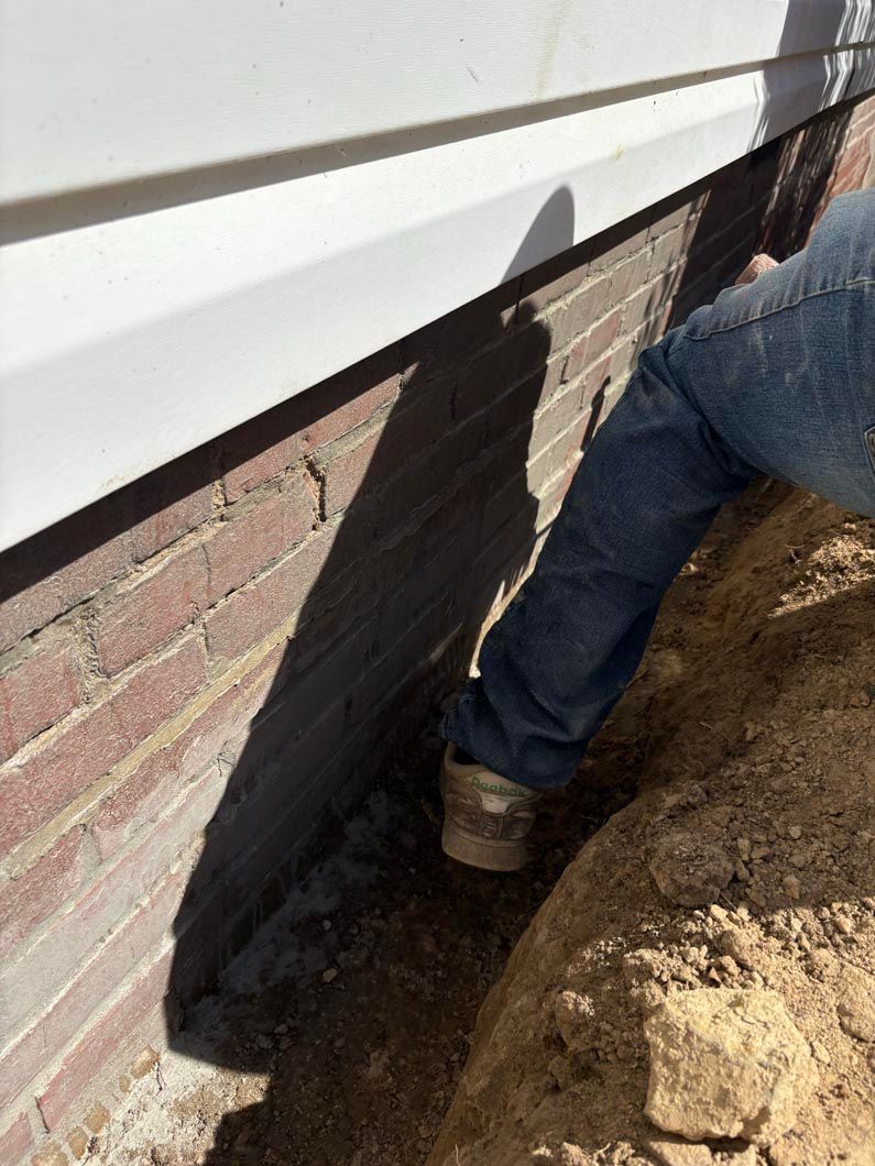 Worker in jeans digging a trench beside a brick house foundation under white siding.