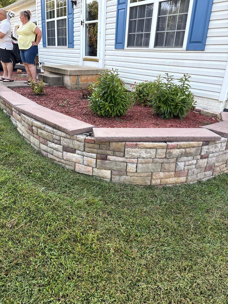 Stone planter bed beside a house with blue shutters; two people work in the garden.