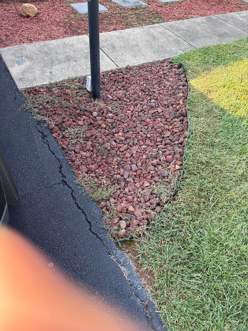Black sidewalk beside red gravel and green grass with a pole and concrete path edge.