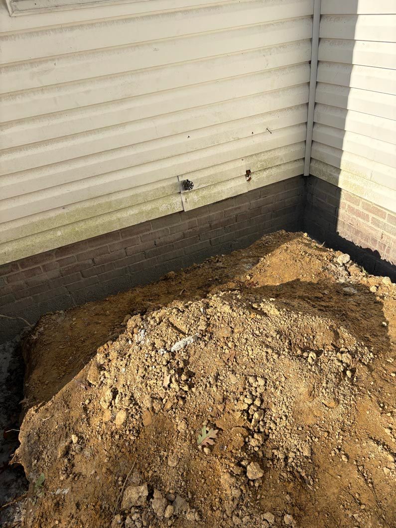 Mound of dirt piled against a house’s foundation beneath cream siding and white brick corner.