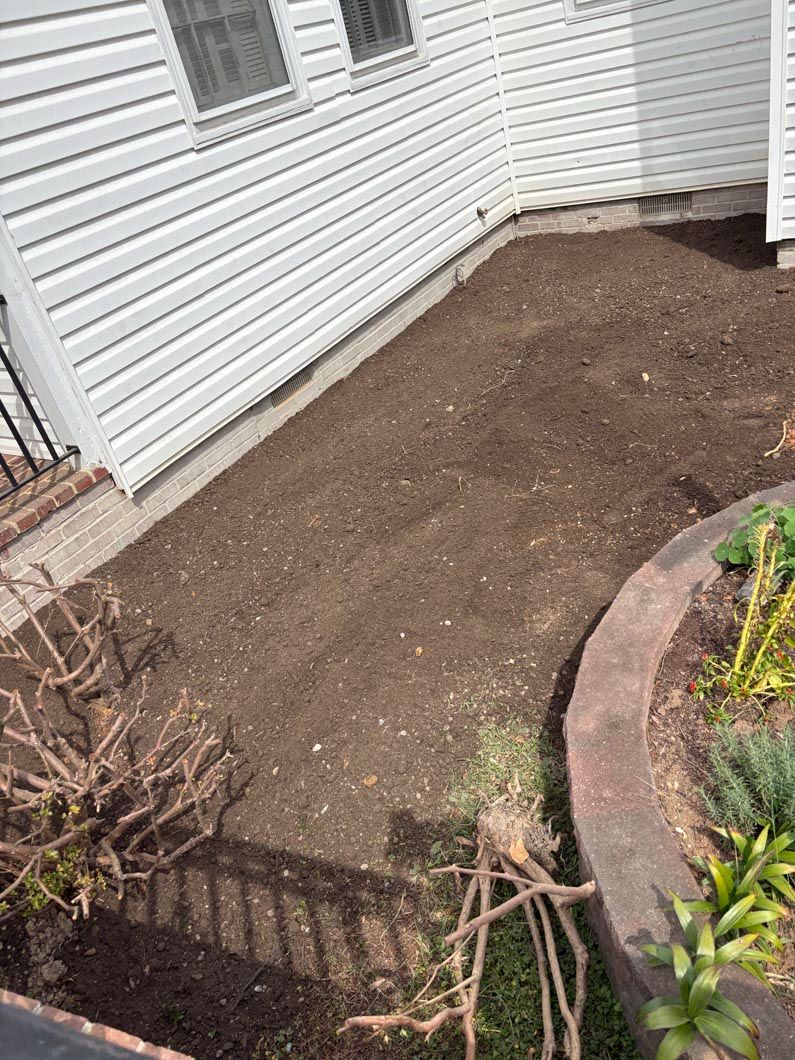 Bare dirt landscaping bed beside a white house, bordered by a curved stone edge and small shrubs.