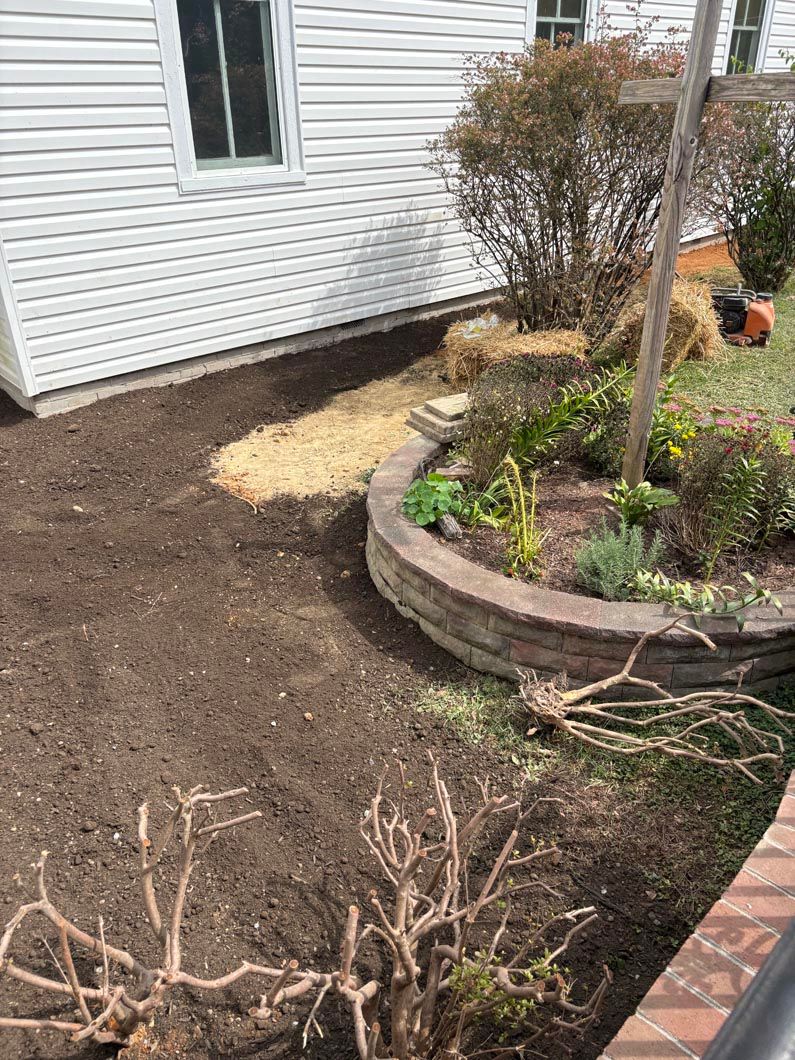 Backyard garden bed with shrubs, fresh mulch, and a white house wall beside a brick walkway.
