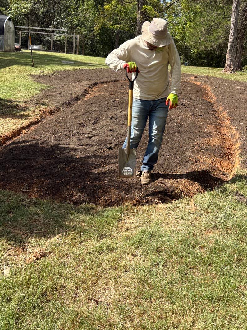 Person spreading mulch along a garden bed with a shovel in a sunny yard