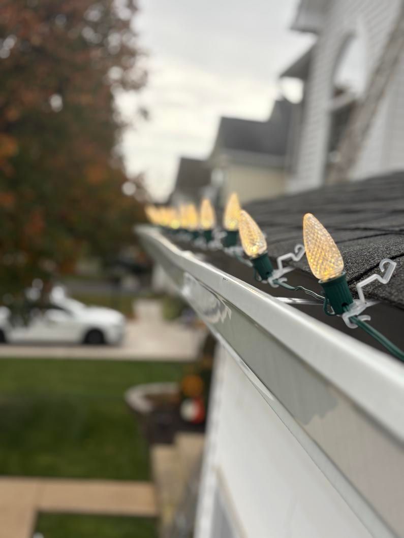 String of warm white holiday lights clipped along a white roof edge, with a blurred house and trees behind.