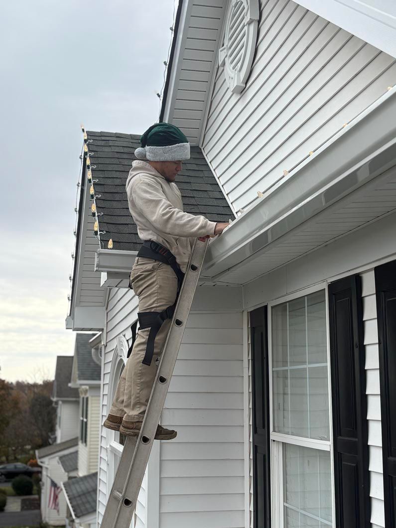 Worker on a ladder cleaning gutters beside a white house.