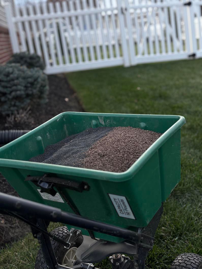 Green spreader cart filled with gravel on a lawn near a white fence.