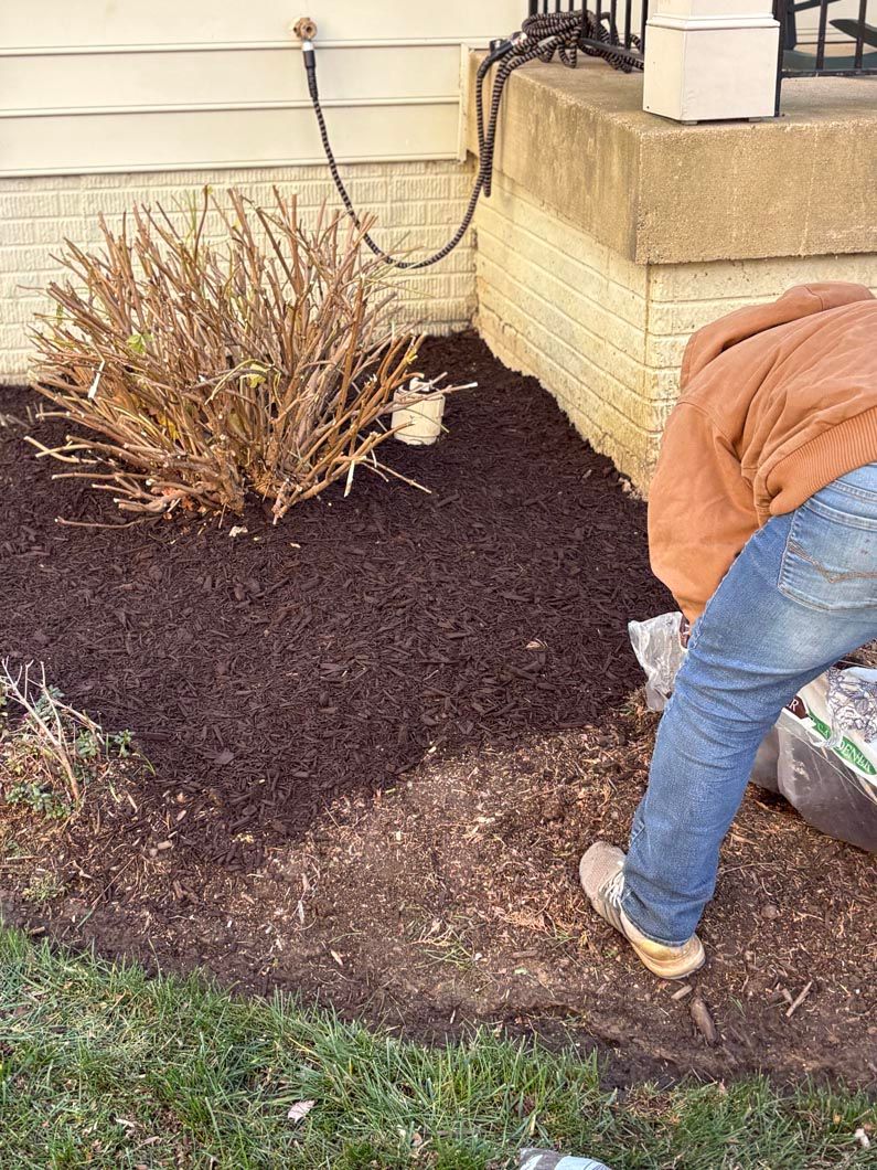 Person edging a garden bed with fresh dark mulch beside a house porch.