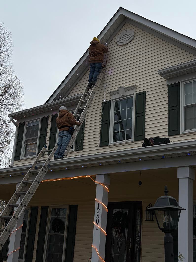 Two workers on ladders repairing the roofline of a white house with green shutters.