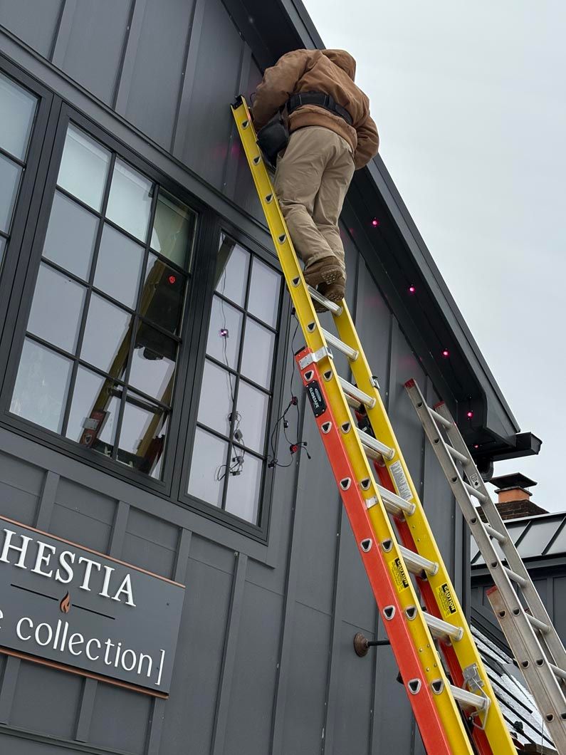 Worker on a yellow ladder repairing a gray storefront roof and windows