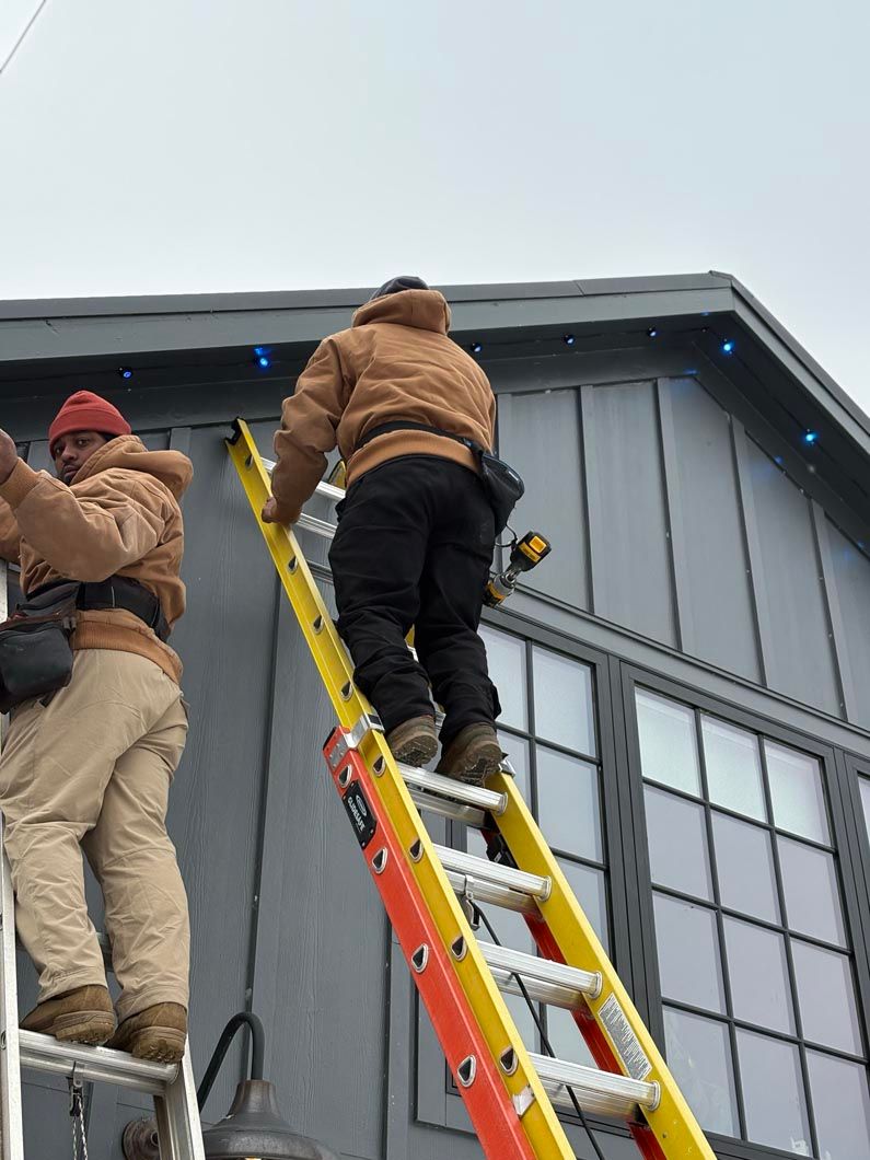 Two workers on ladders repairing the exterior of a gray building near the roofline.
