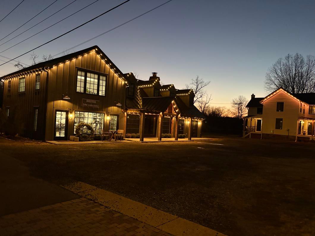 Warmly lit rustic building at dusk with string lights and a neighboring house in the background