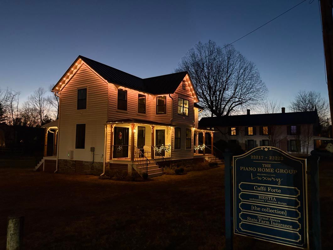 Lit two-story yellow house at dusk with bare trees and a chalkboard sign in front