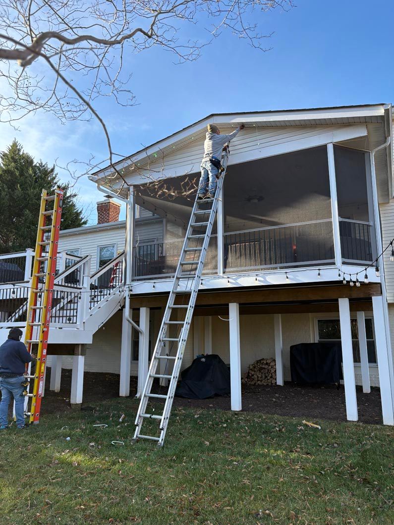 Workers on ladders repairing a raised house’s screened porch and trim outdoors.