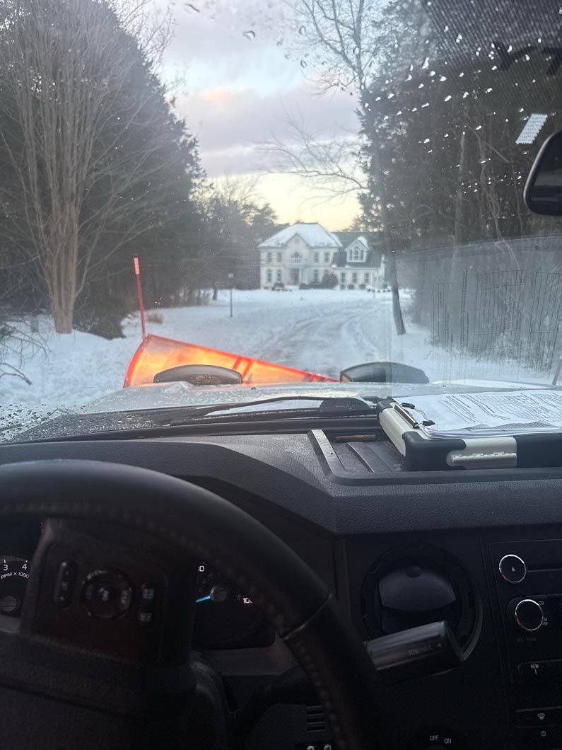 View from a car dashboard on a snowy road, with a tipped orange plow blade ahead near trees and a house.