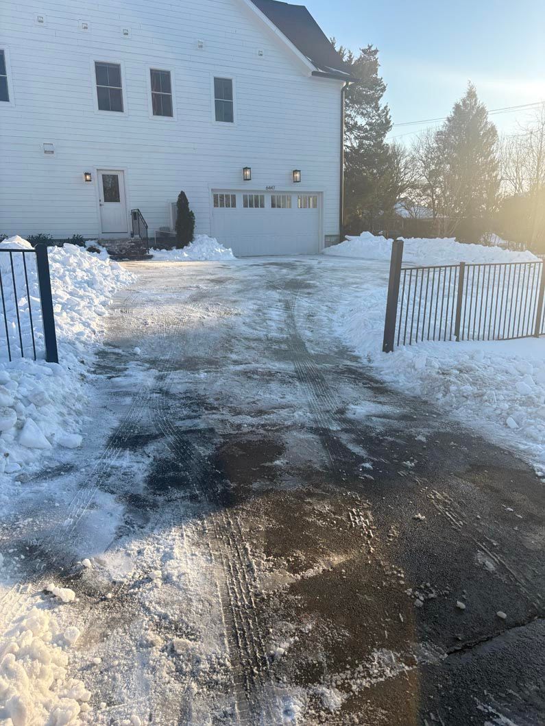 Snow-covered driveway leading to a white house with a fence, lit by low winter sunlight.
