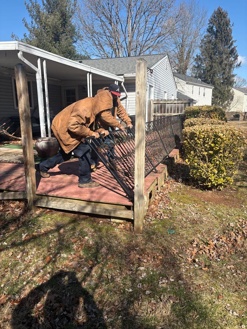 Person in a brown jacket crouches on a porch, working on a wire fence in a sunny backyard.
