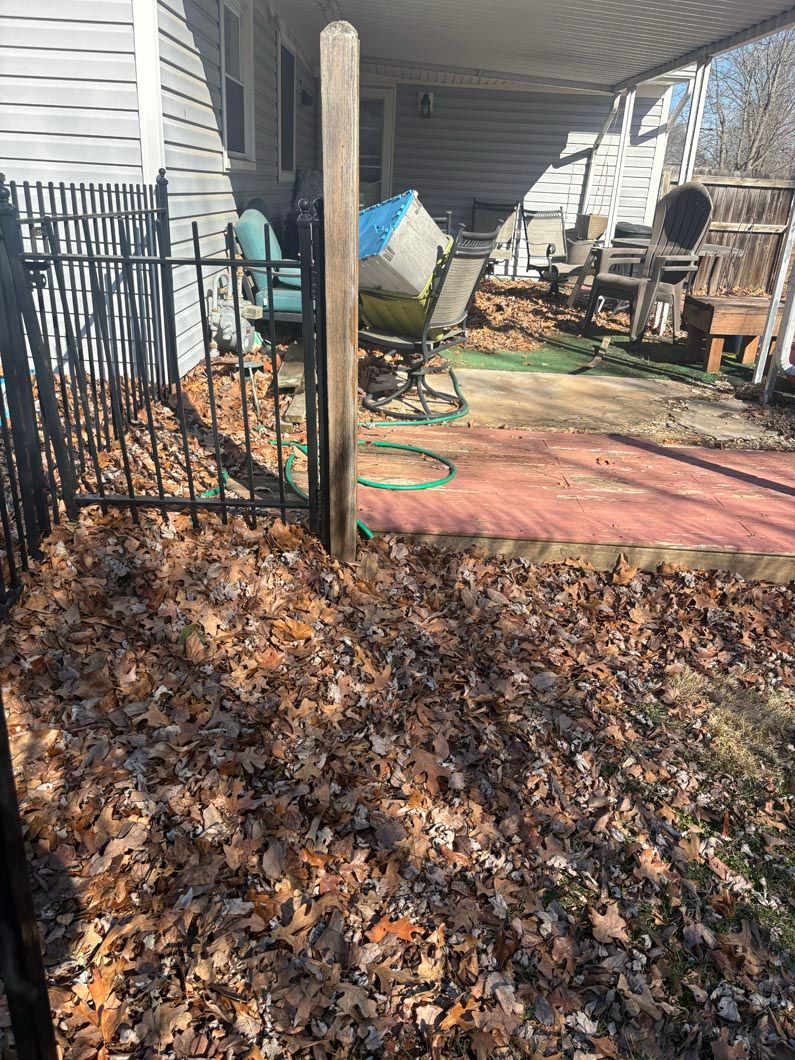 Leaf-covered yard beside a house, with a black fence, patio furniture, and a green hose under a covered porch.
