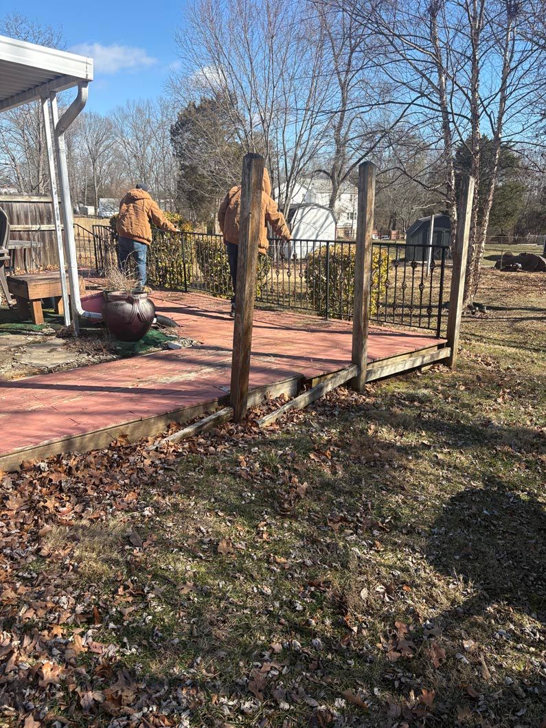 Person in tan coat on a wooden deck with black railing in a leaf-covered yard, near a house on a sunny day
