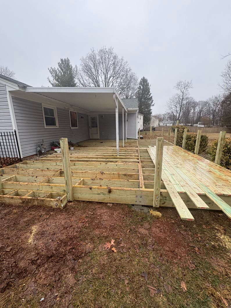 Backyard deck under construction beside a gray house, with wooden framing and planks laid out over muddy ground