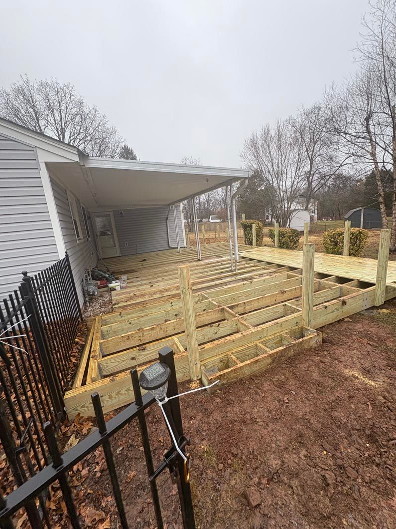 Wooden deck framing under construction beside a gray house, with a covered porch and muddy yard.