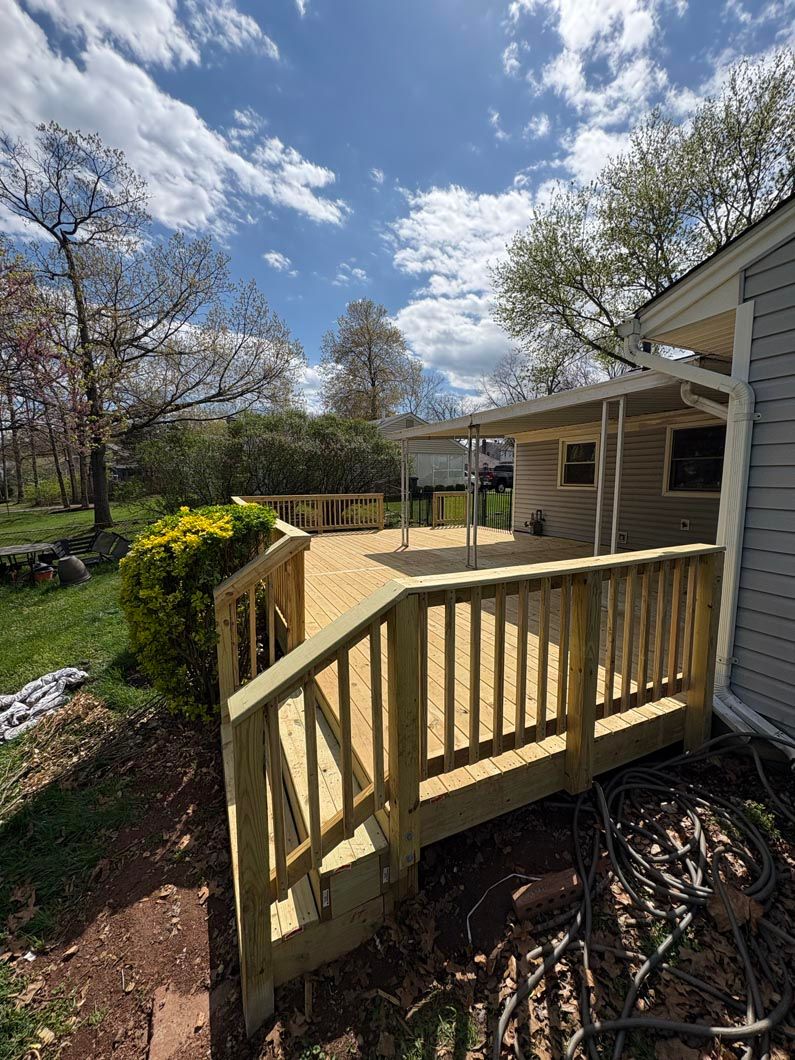 Wooden deck and ramp beside a house, overlooking a sunny garden with trees and flowers.