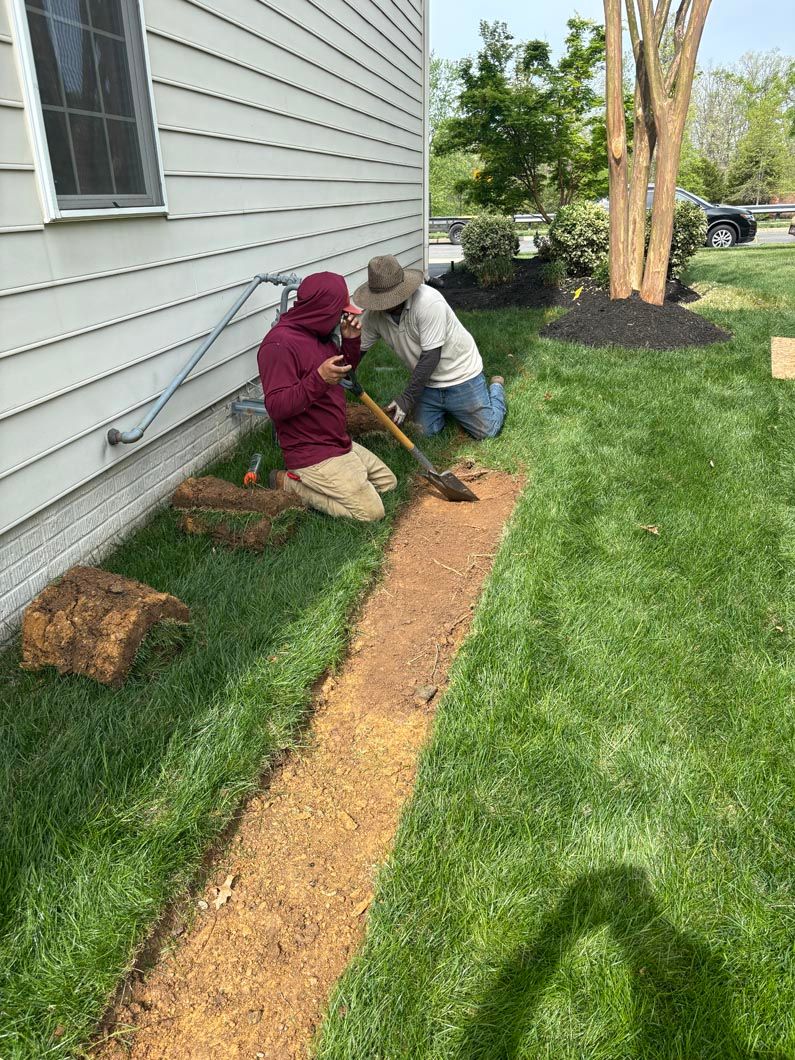 Two people kneel by a house, planting in a garden bed along the siding.
