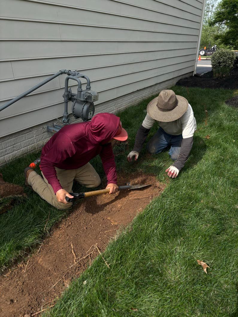 Two people kneel by a house wall, digging a narrow trench along the foundation with tools.