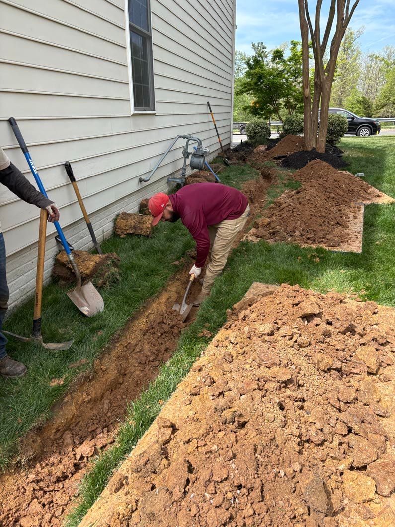 Person digging a narrow trench beside a house, with mulch and garden tools nearby.