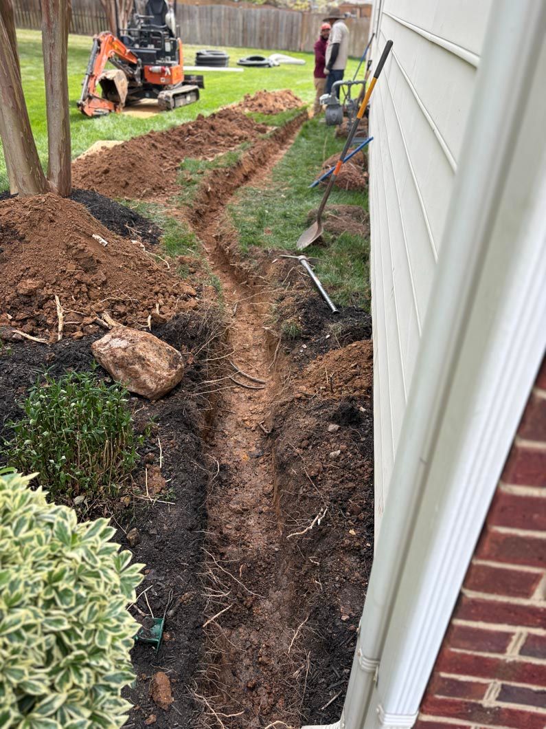 Trench dug beside a house with workers, a small excavator, and uprooted soil along the foundation.