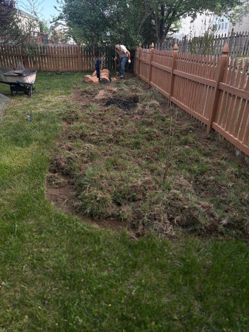 Two people clearing a long pile of yard debris beside a wooden fence in a backyard.
