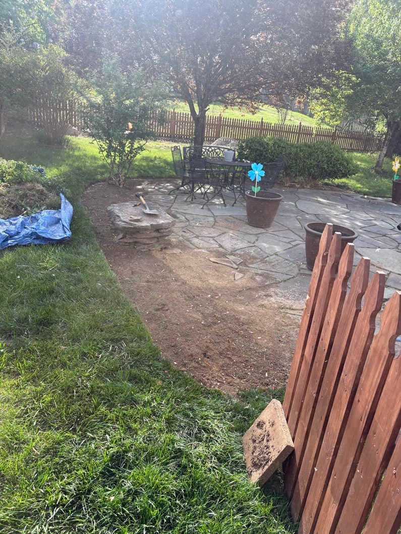 Backyard patio with stone fire pit, potted flowers, grass, and a wooden fence in sunlight