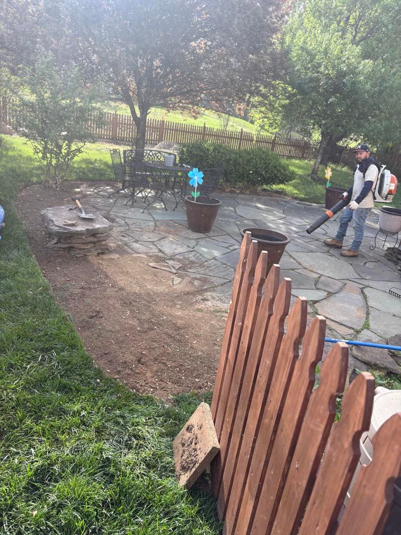 Backyard patio with two people near a stone path, potted plant, and wooden fence in a shaded garden