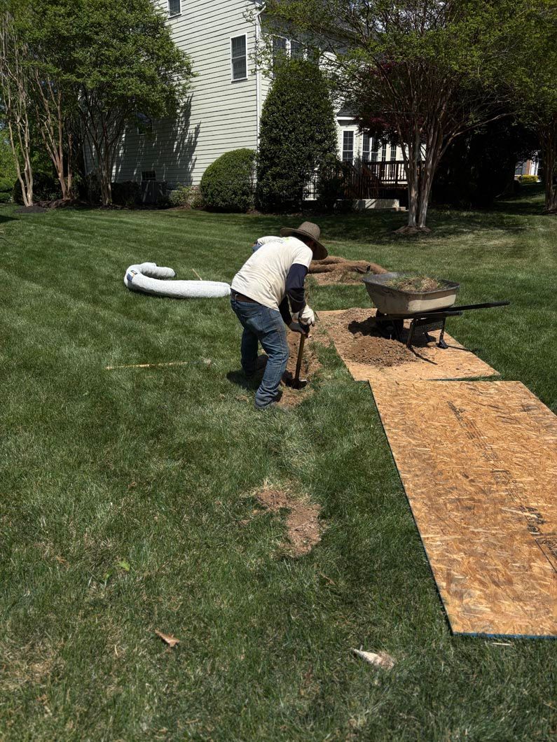 Worker trimming grass near a house, with a white hose and plywood path on the lawn.