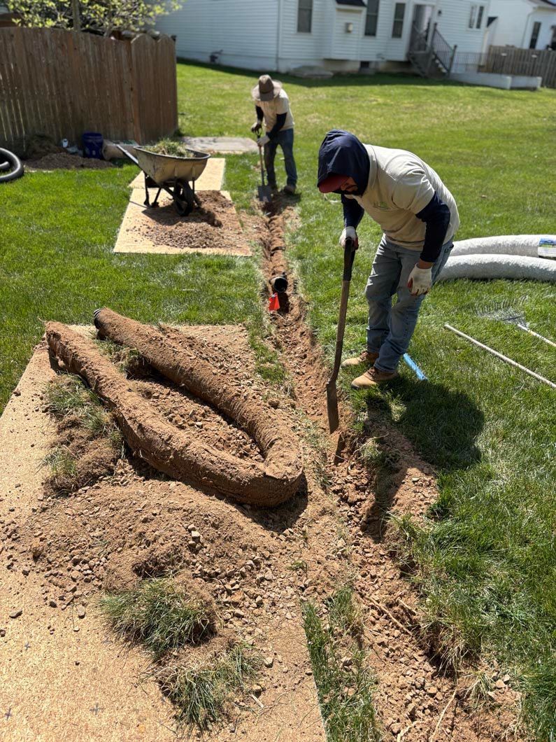 Two workers dig a narrow trench in a grassy yard beside a house, with a wheelbarrow and rolled sod nearby.