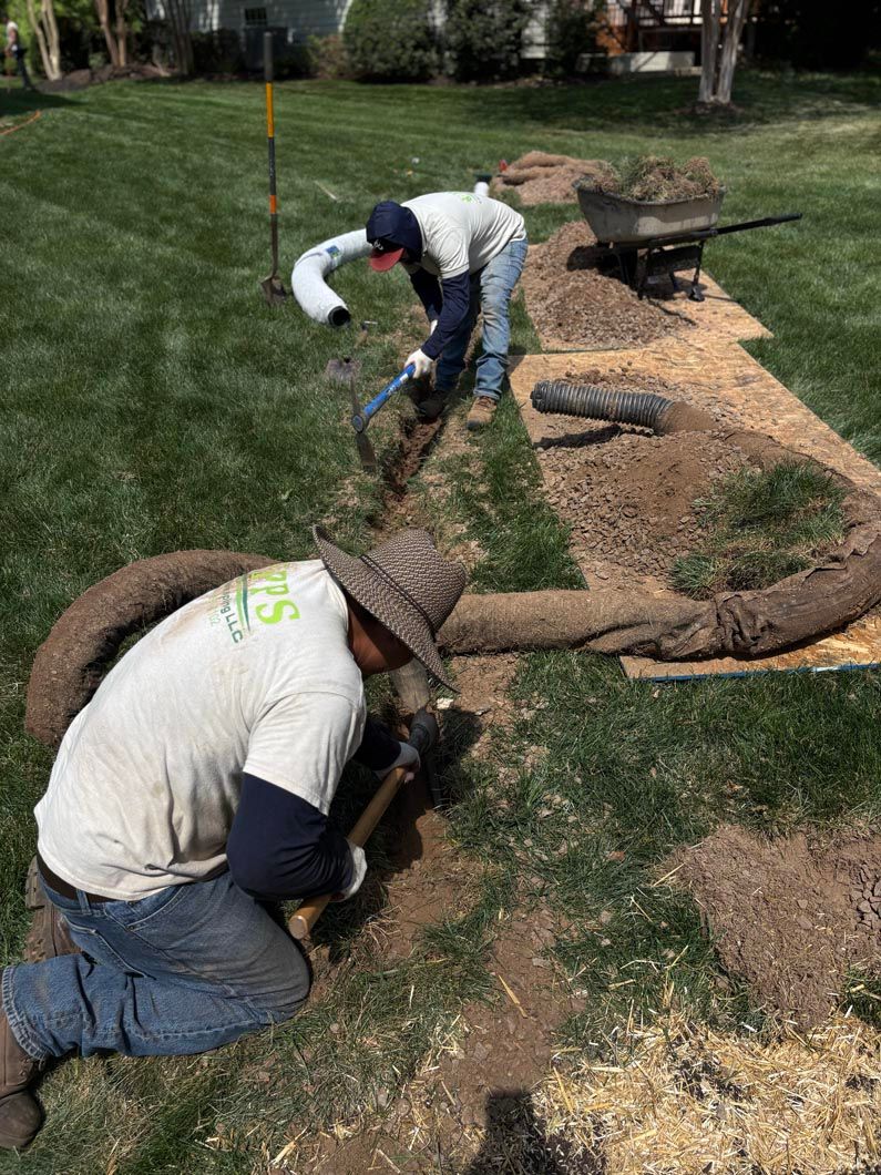 Two workers lay landscape fabric and mulch along a garden bed beside grass.