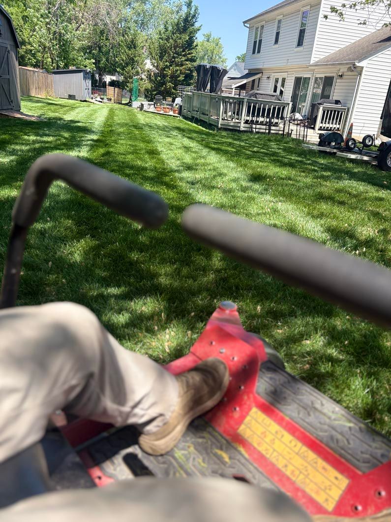 Riding a red lawn mower across a green yard beside a white house and patio chairs