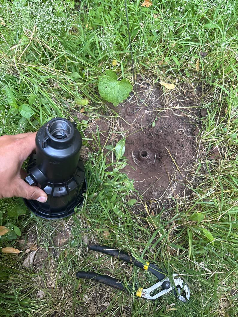 Hand holding a black tool beside a fresh hole in grassy soil, with pliers nearby.