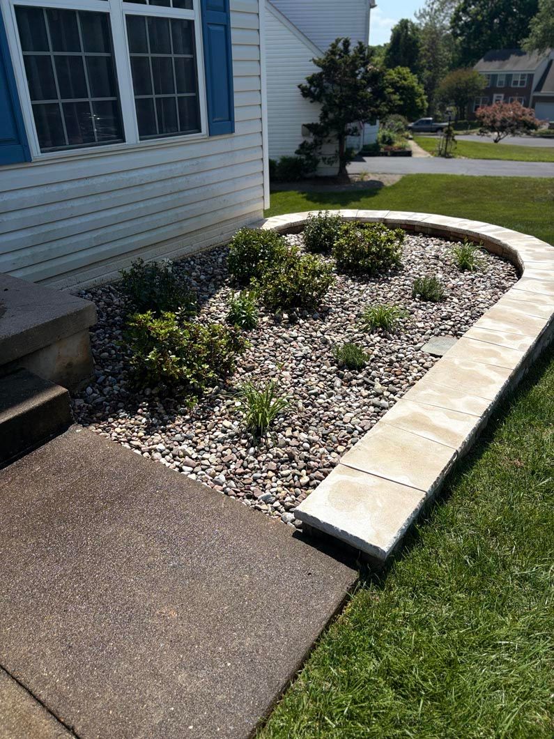 Curved stone-edged garden bed beside a house, with mulch, small plants, a sidewalk, and grass.