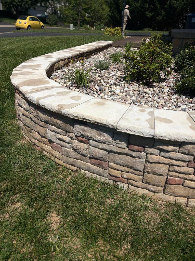 Curved stone retaining wall around a landscaped garden bed with shrubs and white gravel, near a grassy area.