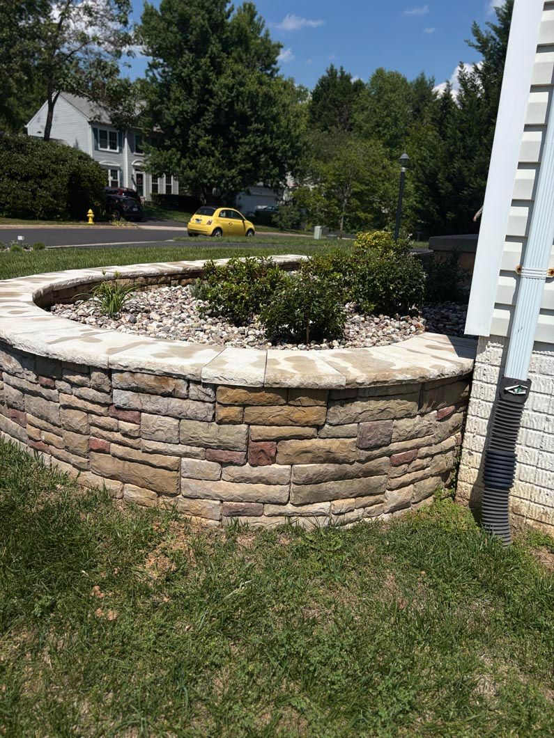 Stone retaining wall around a landscaped circular planter beside a street, with trees and a yellow car in the background
