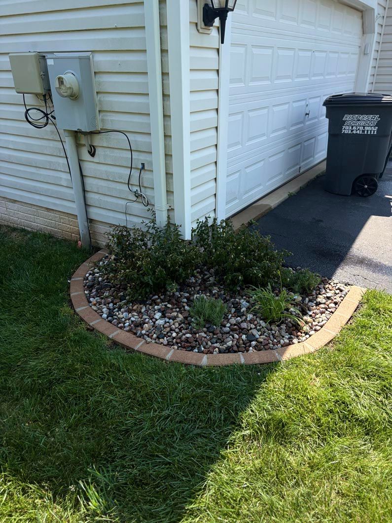 Small landscaped garden bed beside a garage, with green shrubs, mulch, and a black trash bin on the driveway.