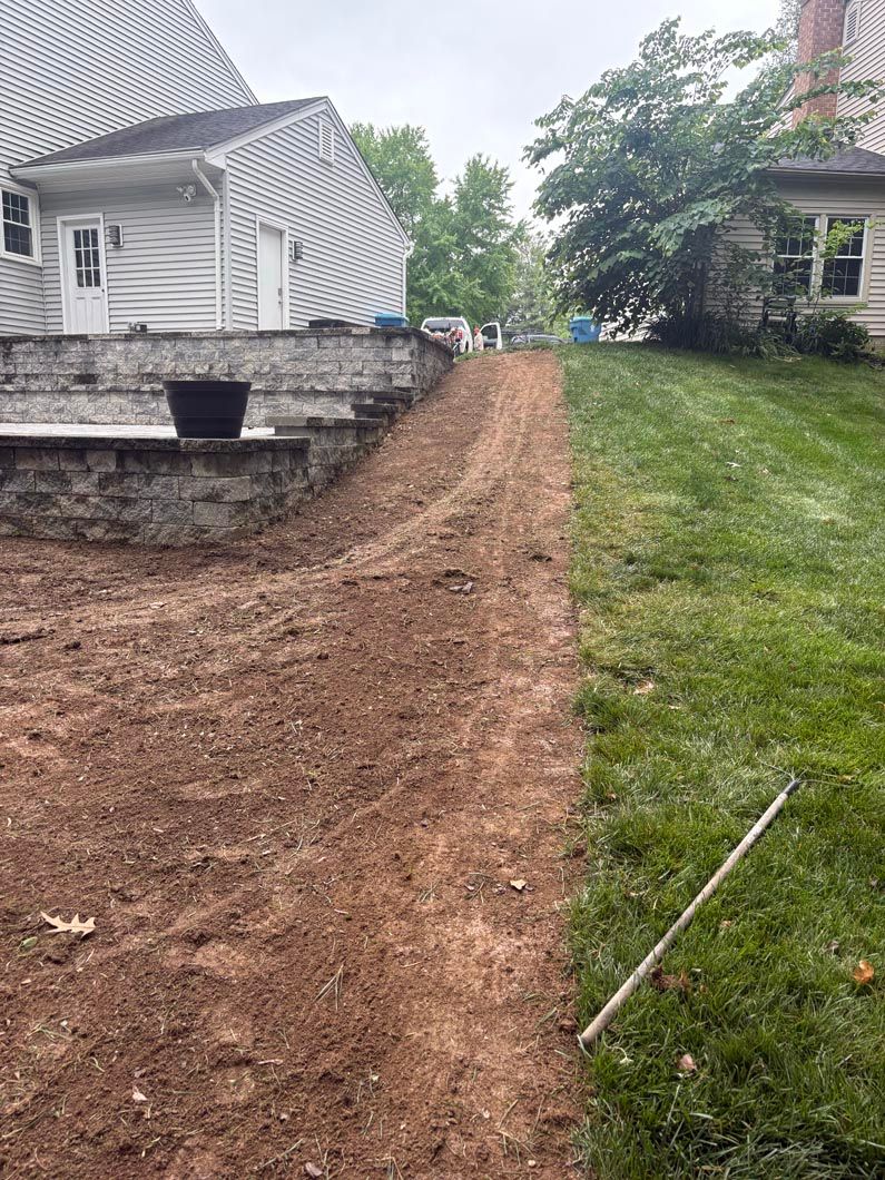 Freshly graded dirt path beside a stone retaining wall between two houses and a grassy yard