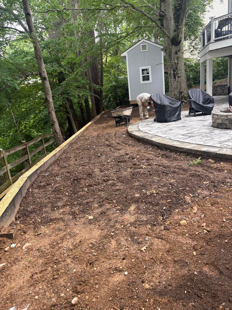 Bare dirt landscaping beside a curved stone patio, with trees and a small shed near a house.