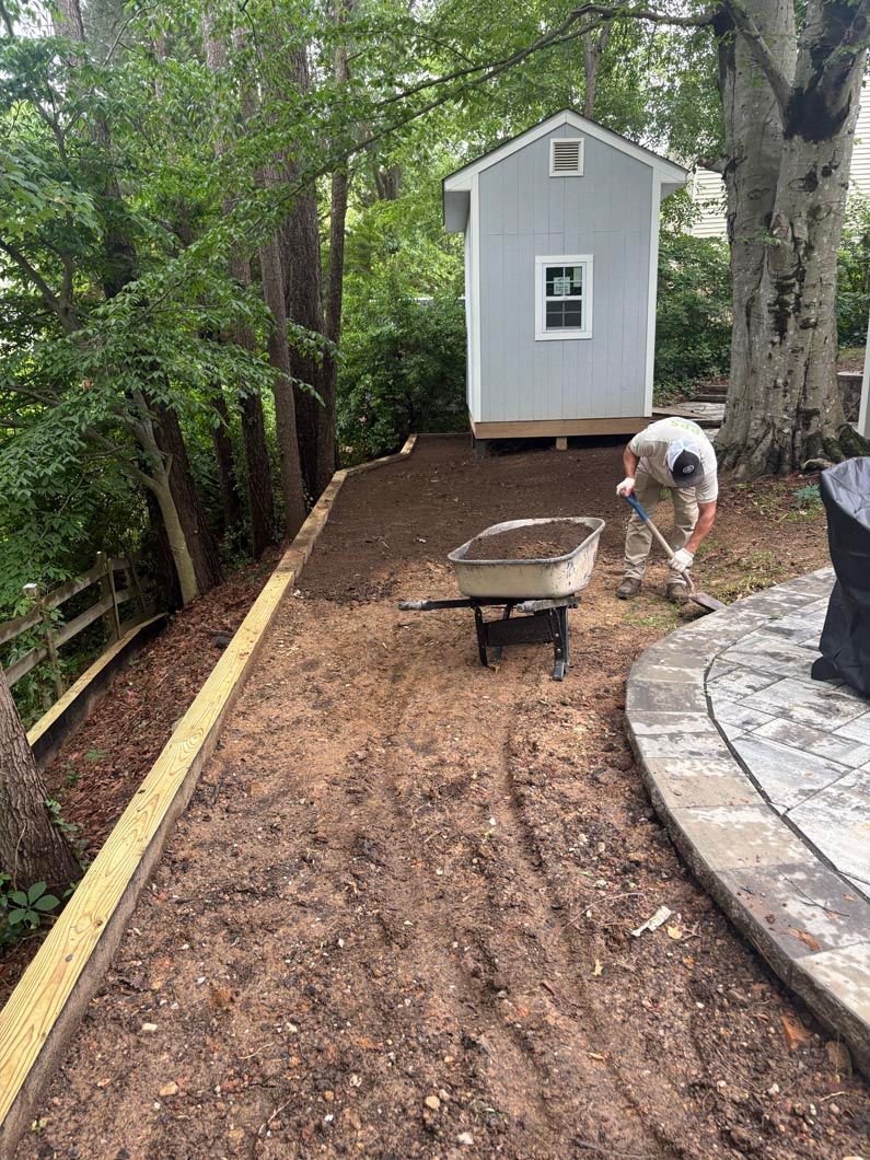 Worker raking mulch on a sloped backyard path beside a patio and shed