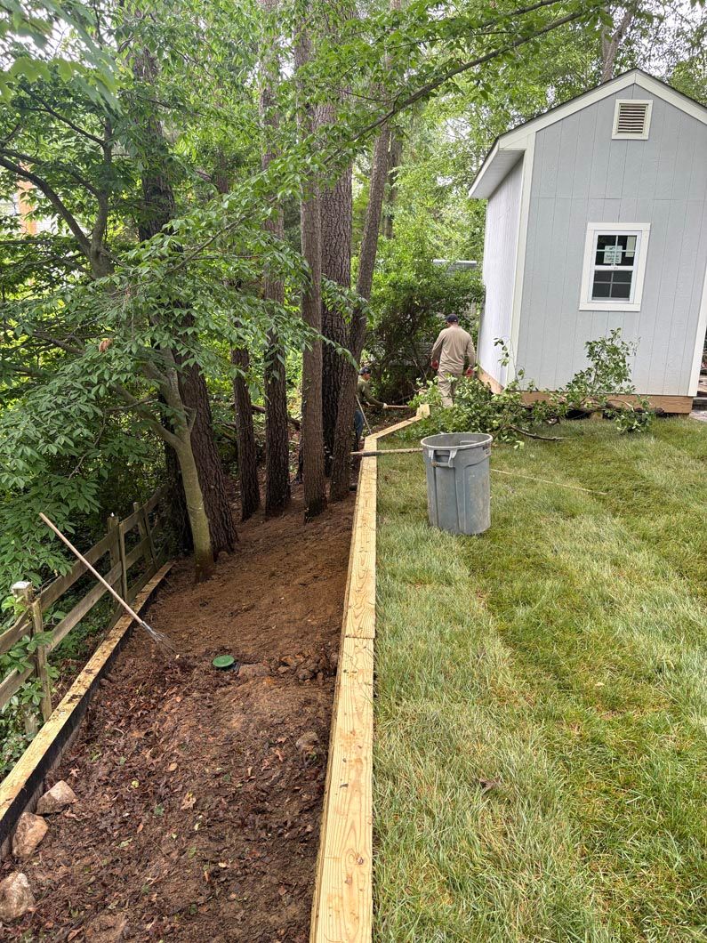 Narrow dirt trench beside a grassy yard and white shed, lined with wood and trees along the edge.