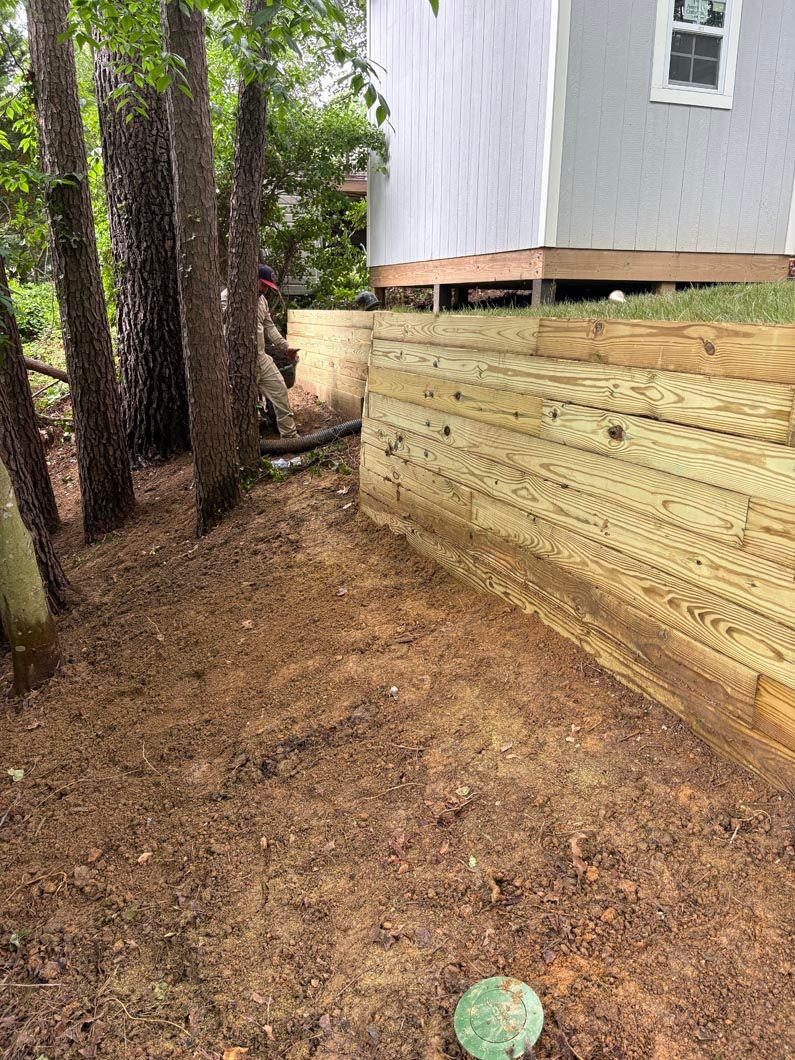 Dirt path beside a raised house with tan retaining wall and trees in a wooded yard