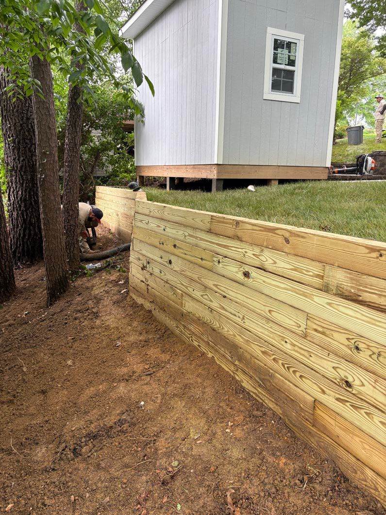 Wooden retaining wall beside a raised white shed, with a tree and dirt path in the foreground.