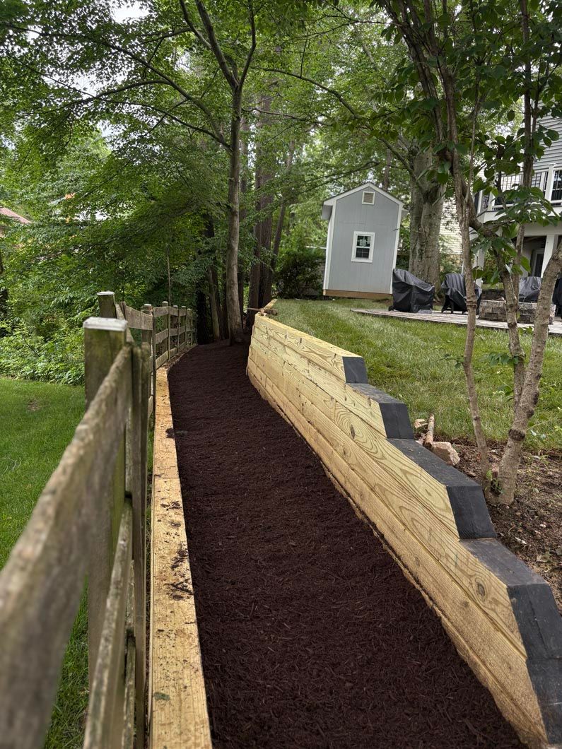 Curved mulch path beside a stone retaining wall, leading past trees to a small shed in a backyard.