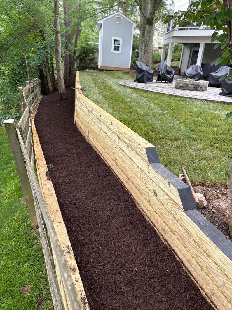 Narrow mulched side yard path beside a retaining wall and lawn, with a shed and patio chairs in the background