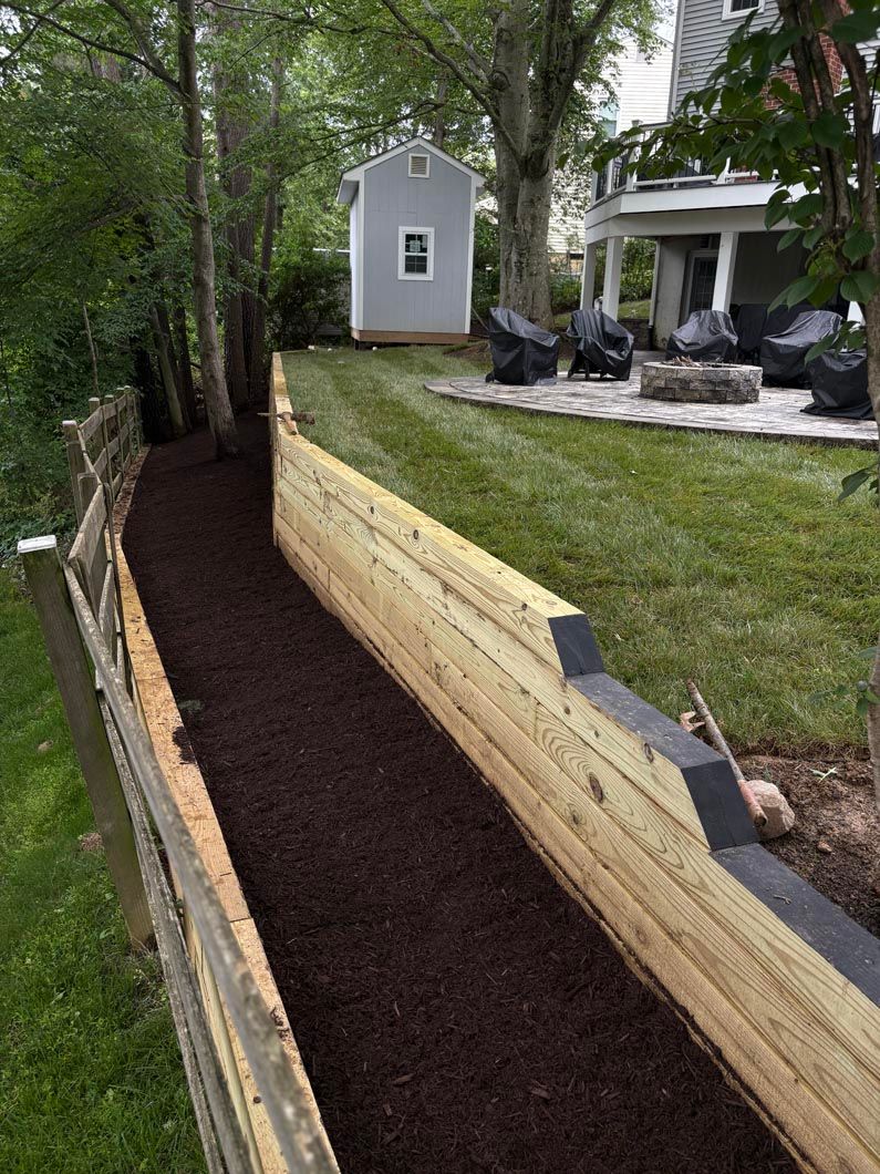 Wooden retaining wall beside a mulched walkway and grassy yard with a small shed and patio chairs.